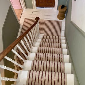 May include: Staircase with white risers and a runner with a beige background and vertical burgundy stripes. The handrail is wood with white balusters. A pink door is visible at the top of the stairs.