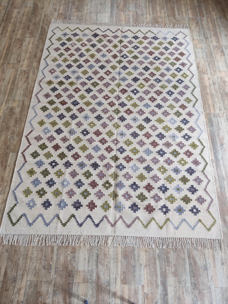 May include: A white rug with a geometric pattern in shades of green, purple, and blue. The rug has a fringe border.