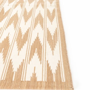 Puede incluir: Una alfombra tejida en color beige y blanco con un patr&oacute;n de chevron. La alfombra tiene un borde de flecos.