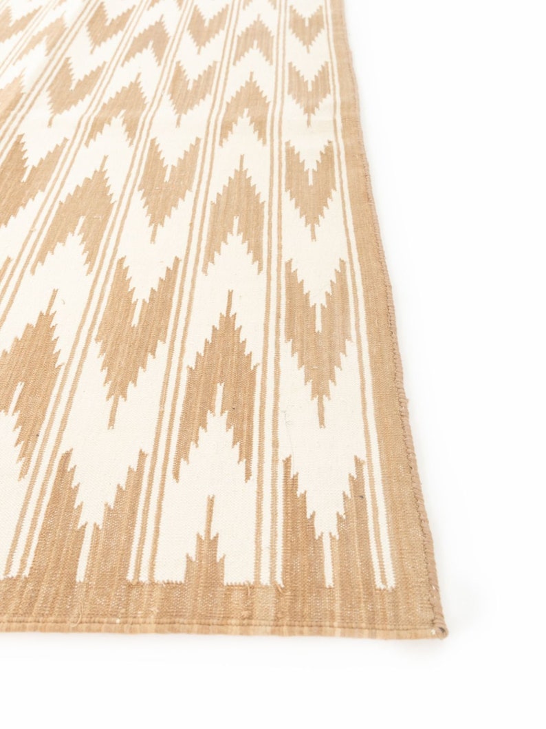 Puede incluir: Una alfombra tejida en color beige y blanco con un patr&oacute;n de chevron. La alfombra tiene un borde de flecos.