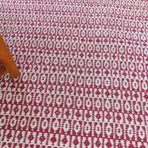 May include: A close-up of a red and white patterned rug. The rug has a repeating geometric design with diamond and rectangular shapes. The color scheme is a bold red and white, creating a striking visual contrast. The rug appears to be made of woven material.