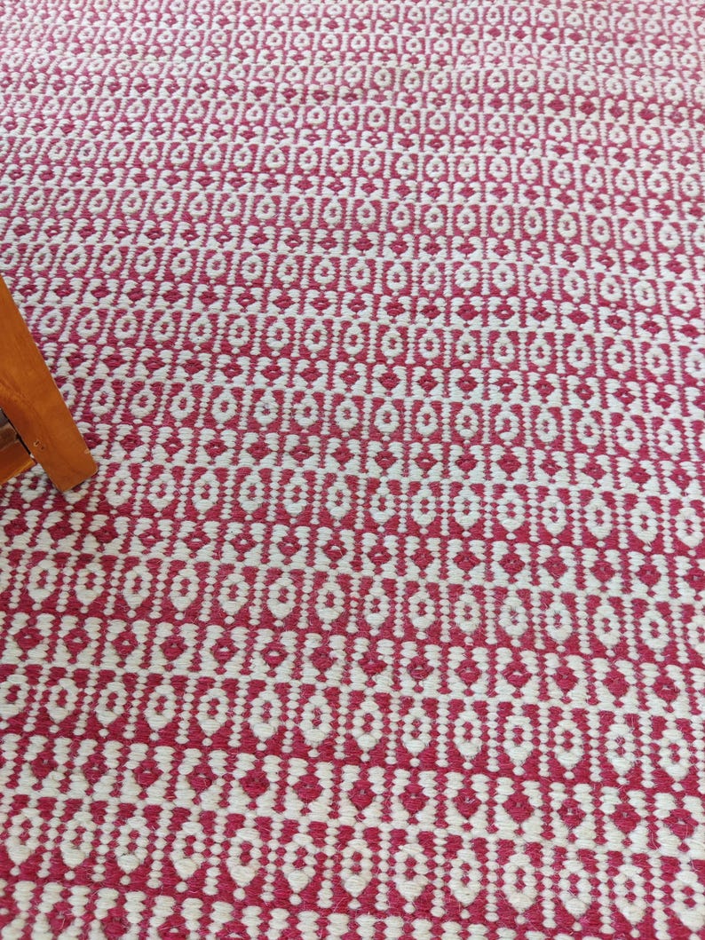 May include: A close-up of a red and white patterned rug. The rug has a repeating geometric design with diamond and rectangular shapes. The color scheme is a bold red and white, creating a striking visual contrast. The rug appears to be made of woven material.