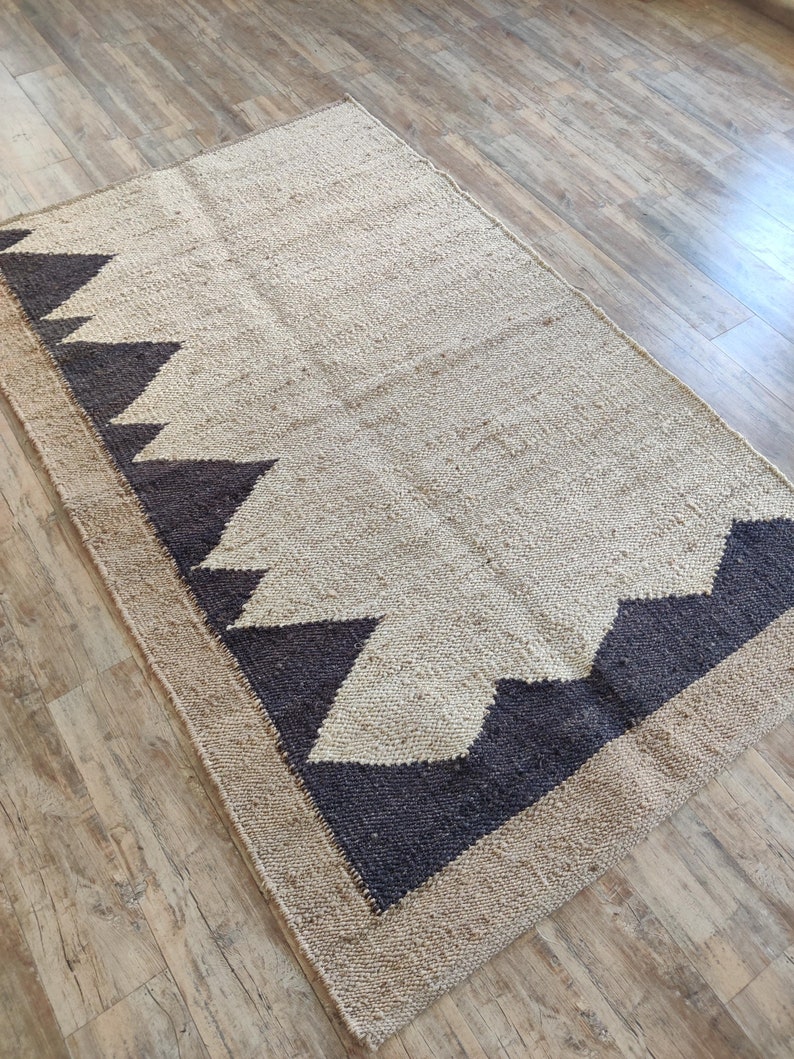 K&ouml;nnte beinhalten: Ein beige-schwarzer Teppich mit geometrischem Muster. Der Teppich hat eine gewebte Textur und einen Fransenrand.