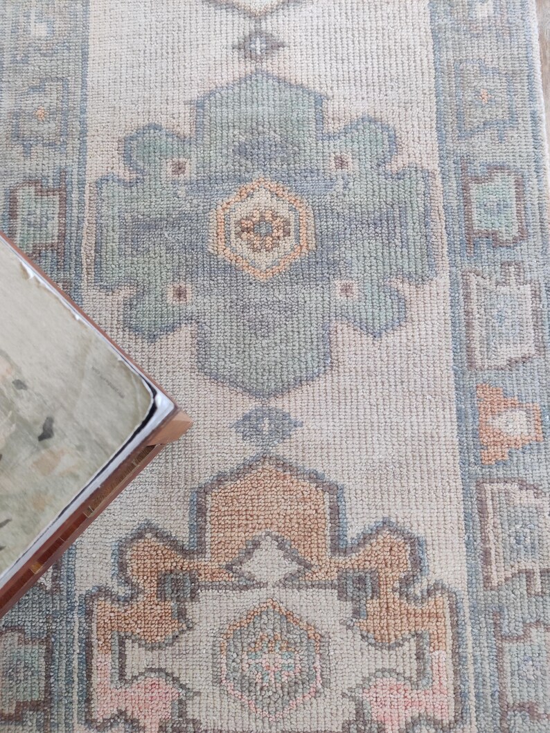 May include: A light beige rug with a geometric pattern in shades of blue, green, and pink. The rug has a fringed border.