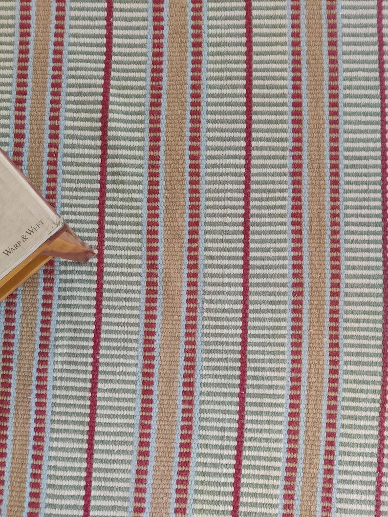 handloom wool blanket showcasing a vibrant striped pattern in red, green, and beige. The wool texture appears soft and inviting, ideal for home decor use. This woolen throw adds a cozy touch to any living room or bedroom, epitomizing luxury textiles with its premium craftsmanship. Known for its durability and softness, this artisan blanket suits all seasons and enhances any space with its elegant desig