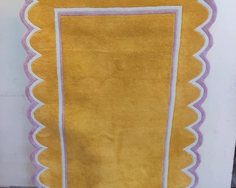 May include: A rectangular rug with a scalloped edge. The rug features a mustard yellow center, framed by a white and light purple border. The outer edge is a scalloped light purple. The rug is made of a soft material.