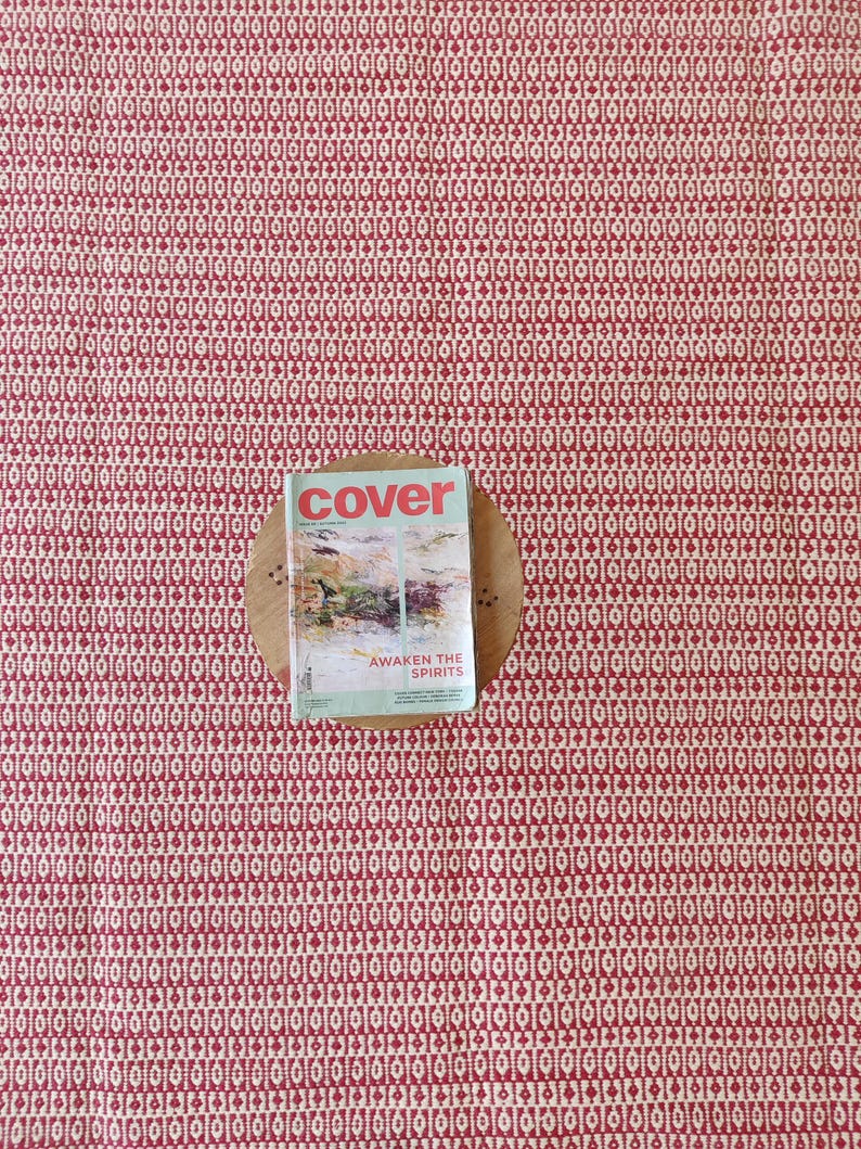 May include: A red and cream-colored patterned rug with a magazine on a wooden coaster. The magazine cover reads "cover" with the text "AWAKEN THE SPIRITS" below a landscape image. The rug's pattern is a repeating geometric design.