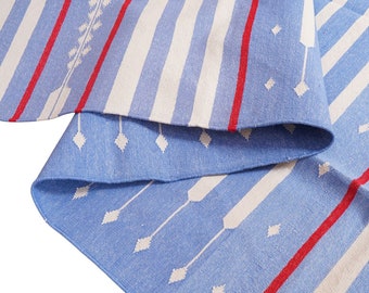 May include: A blue and white striped rug with red accents and white arrow and diamond patterns. The rug has a fringed edge and is folded over, showcasing the design. The rug is made of a woven material.
