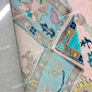 K&ouml;nnte beinhalten: Nahaufnahme eines Teils eines rosa und t&uuml;rkisfarbenen Teppichs mit floralen und geometrischen Mustern. Der Teppich hat einen hellbeigen Rand und der Text "HOUSE OF KILIMS" ist sichtbar.
