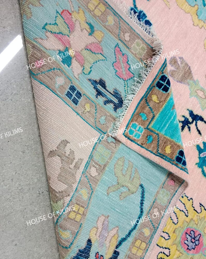 K&ouml;nnte beinhalten: Nahaufnahme eines Teils eines rosa und t&uuml;rkisfarbenen Teppichs mit floralen und geometrischen Mustern. Der Teppich hat einen hellbeigen Rand und der Text "HOUSE OF KILIMS" ist sichtbar.