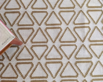May include: Cream-colored rug with a repeating geometric pattern of brown triangles. The triangles are outlined in brown and filled with the cream color of the rug. The rug has a soft, textured appearance. A book with the text "Warp & Weft" is partially visible in the corner.