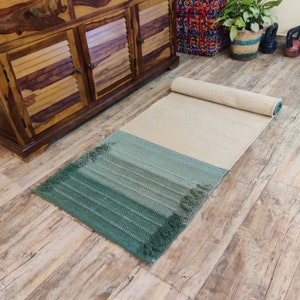 May include: A green and beige woven rug with fringed edges. The rug is rolled up and has a light beige color on the top half and a darker green color on the bottom half.