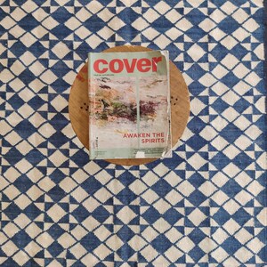 Pu&ograve; includere: Una rivista intitolata "cover" con scritte rosse, appoggiata su un tavolino di legno. La rivista presenta un dipinto astratto e il testo "AWAKEN THE SPIRITS". Il tavolo &egrave; su un tappeto a motivi blu e bianchi.