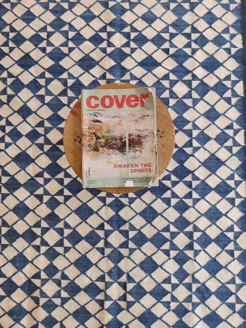 Pu&ograve; includere: Una rivista intitolata "cover" con scritte rosse, appoggiata su un tavolino di legno. La rivista presenta un dipinto astratto e il testo "AWAKEN THE SPIRITS". Il tavolo &egrave; su un tappeto a motivi blu e bianchi.