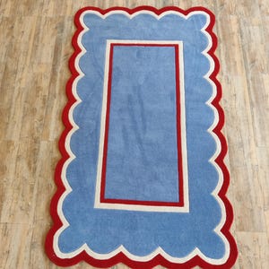 May include: A rectangular rug with a scalloped edge. The rug features a light blue center, a white border, and a red outer edge. The rug's design is reminiscent of a picture frame.