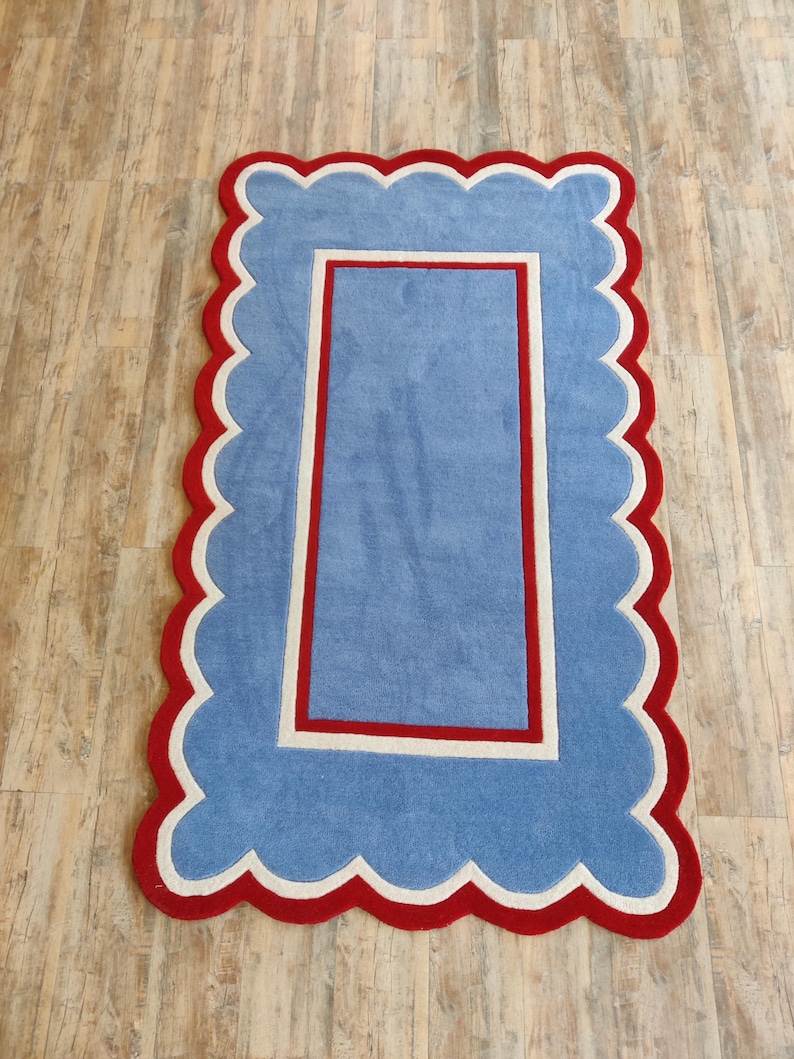 May include: A rectangular rug with a scalloped edge. The rug features a light blue center, a white border, and a red outer edge. The rug's design is reminiscent of a picture frame.