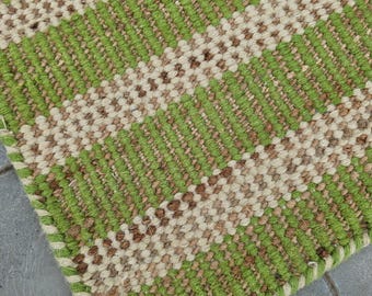 May include: A green and beige striped woven rug with a fringed edge. The rug is made of natural fibers and has a rustic look.