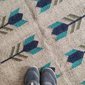 Op de afbeelding: Een paar grijze sportschoenen met zwarte veters en witte accenten op een tapijt met een patroon van blauwe, groene en bruine pijlen.