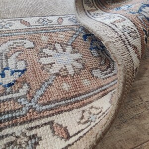 May include: Close-up of the edge of a beige rug with a brown and blue patterned border. The rug has a soft, woven texture.