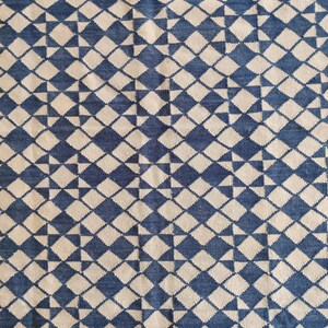 Pu&ograve; includere: Primo piano di un tessuto con un motivo geometrico ripetuto. Il design presenta forme a diamante blu e bianco sporco alternate, creando una superficie strutturata. Il tessuto sembra essere tessuto, con una trama visibile.