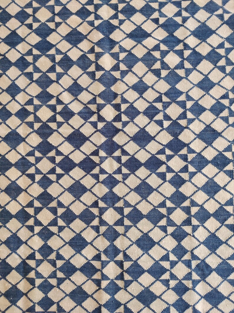 Pu&ograve; includere: Primo piano di un tessuto con un motivo geometrico ripetuto. Il design presenta forme a diamante blu e bianco sporco alternate, creando una superficie strutturata. Il tessuto sembra essere tessuto, con una trama visibile.