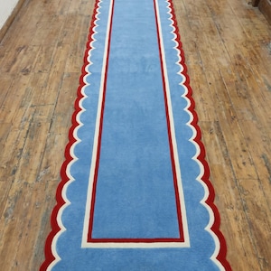 May include: A rectangular blue rug with a scalloped red border and a white and red inner border. The rug is placed on a wooden floor. The rug's design is simple and elegant, suitable for a hallway or entryway.