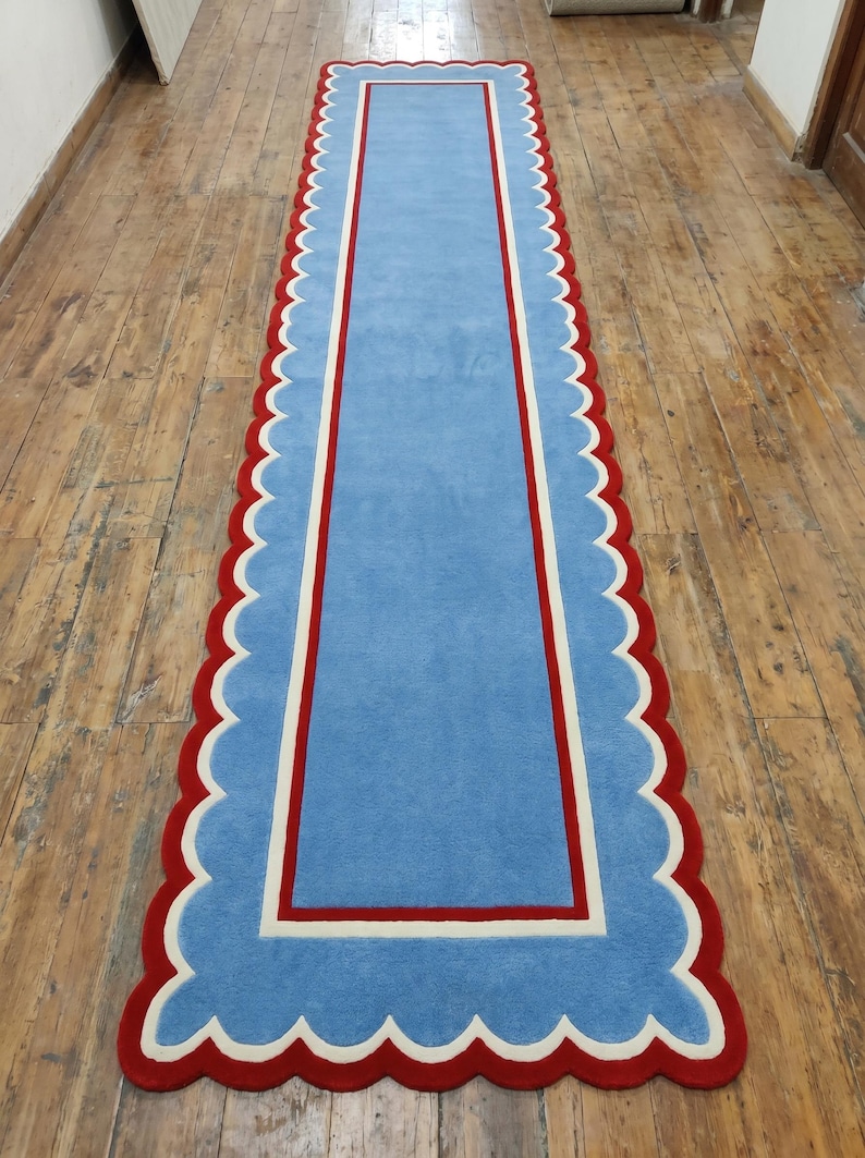May include: A rectangular blue rug with a scalloped red border and a white and red inner border. The rug is placed on a wooden floor. The rug's design is simple and elegant, suitable for a hallway or entryway.