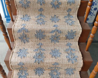 May include: A jute stair runner in natural tones, featuring a repeating blue floral design. The runner is laid on wooden stairs with wooden banisters. The pattern provides a decorative touch to the staircase.