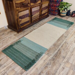 May include: A green and cream woven rug with fringed edges. The rug is laid out on a wooden floor.