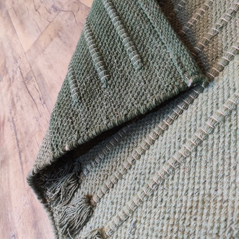 May include: A green woven rug with a textured pattern. The rug has a fringe border.