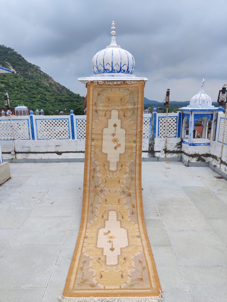 May include: A long, beige runner rug with a traditional floral pattern. The rug is displayed on a rooftop with a blue and white dome in the background.