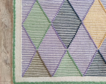 May include: A woven rug with a geometric pattern of diamonds in shades of purple, green, yellow, and blue. The rug has a white fringe border.