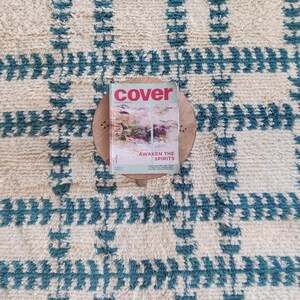 May include: A white and blue patterned rug with a magazine titled "cover" on top of a wooden round stand. The magazine cover features a colorful abstract design and the text "Awaken the Spirits".