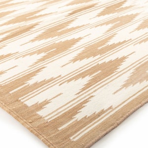 Puede incluir: Una alfombra tejida beige y blanca con un patr&oacute;n geom&eacute;trico. La alfombra tiene un borde con flecos.