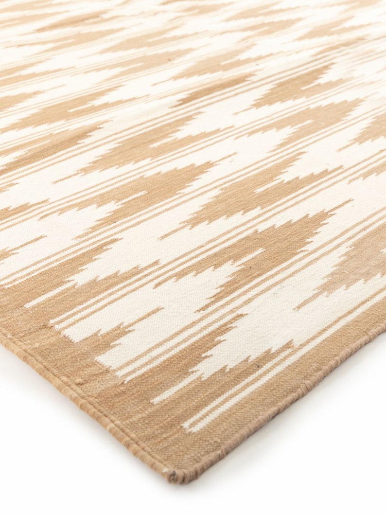 Puede incluir: Una alfombra tejida beige y blanca con un patr&oacute;n geom&eacute;trico. La alfombra tiene un borde con flecos.