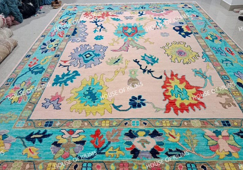K&ouml;nnte beinhalten: Ein gro&szlig;er, rechteckiger Teppich mit einem hellrosa Mittelfeld und einem t&uuml;rkisfarbenen Rand. Der Teppich weist farbenfrohe Blumenmuster in Blau-, Gelb-, Rot- und Gr&uuml;nt&ouml;nen auf. Der Text "HOUSE OF KILIMS" ist am Rand sichtbar.