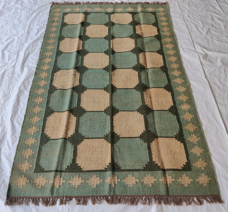 May include: A green and beige patterned rug with a geometric design. The rug has a fringe border.