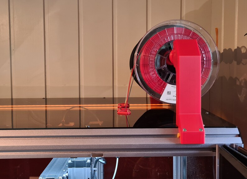 May include: A red filament spool holder attached to a 3D printer. The spool is clear and contains red filament.