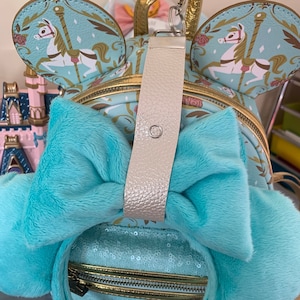 May include: A turquoise blue faux fur Minnie Mouse headband with a gold zipper and a white bow. The headband is decorated with sequins and has a gold chain attached to the bow.