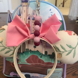 May include: A white headband with pink fabric bow and a purple and pink beaded charm. The headband is in front of a blue and purple castle themed Loungefly backpack.