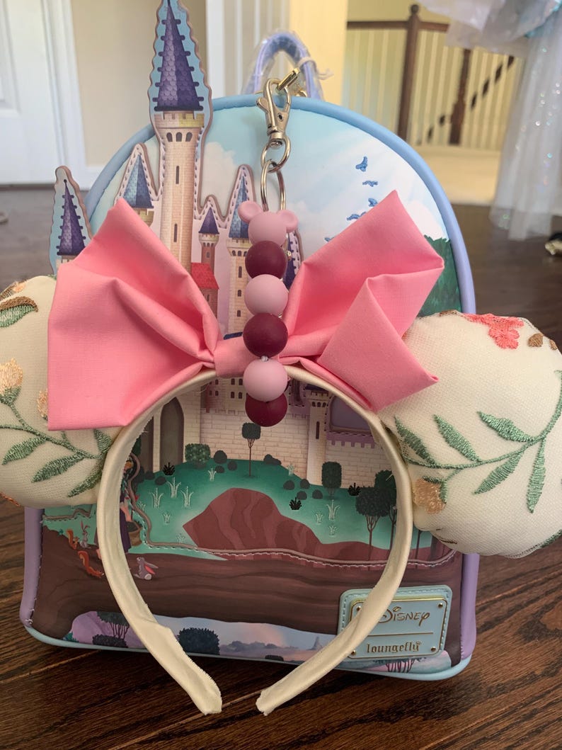 May include: A white headband with pink fabric bow and a purple and pink beaded charm. The headband is in front of a blue and purple castle themed Loungefly backpack.