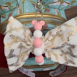 May include: A white and gold bow with a pink and white beaded keychain attached. The keychain is attached to a silver key ring.