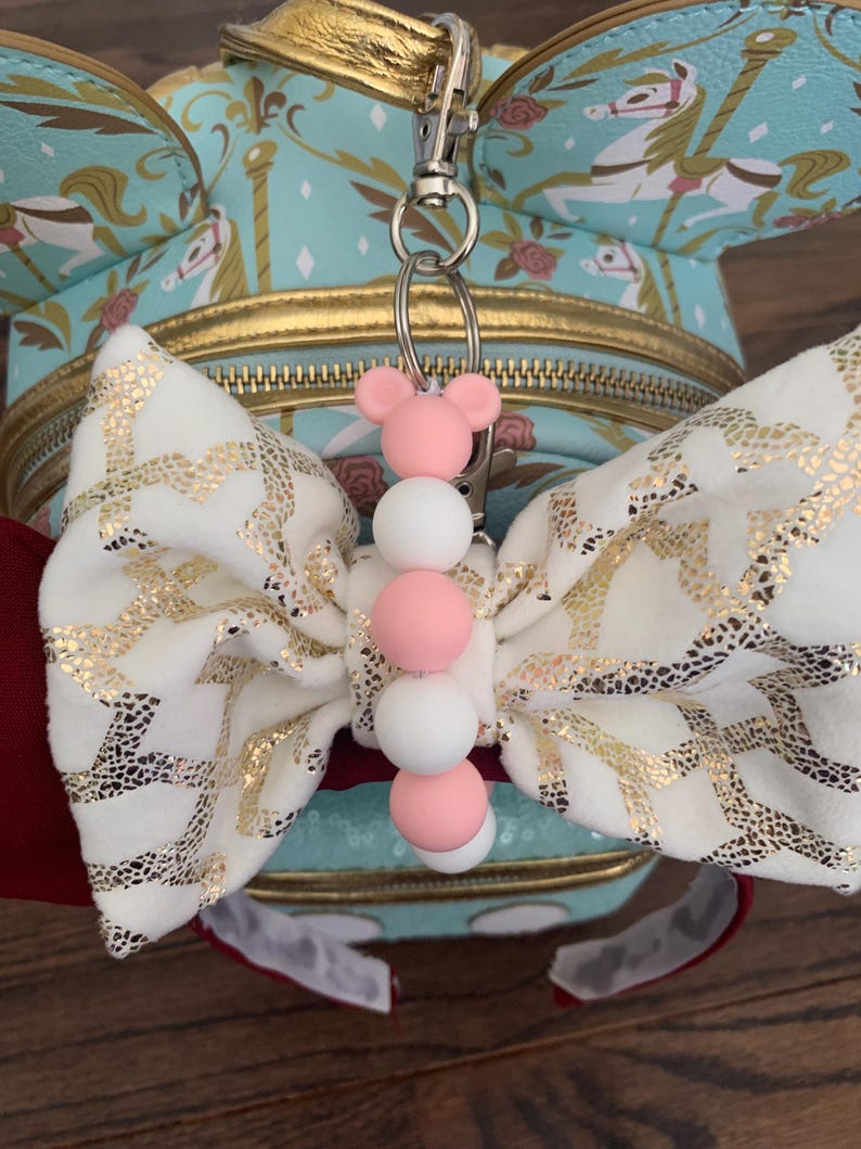 May include: A white and gold bow with a pink and white beaded keychain attached. The keychain is attached to a silver key ring.