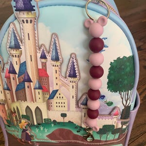 May include: A light blue backpack with a castle illustration and a purple, pink, and burgundy beaded keychain with a Mickey Mouse ear charm. The backpack has a zipper and a Disney label that says "Loungefly".