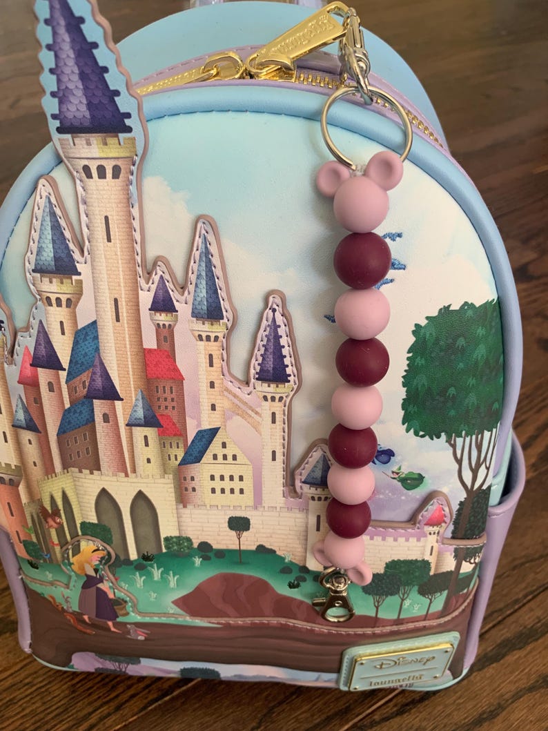May include: A light blue backpack with a castle illustration and a purple, pink, and burgundy beaded keychain with a Mickey Mouse ear charm. The backpack has a zipper and a Disney label that says "Loungefly".