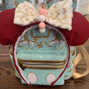May include: A turquoise blue and gold mini backpack with a white and gold bow, red ears, and a white and pink charm. The backpack has a zippered pocket and a gold chain. The backpack has the text "Disney Loungefly" on it.