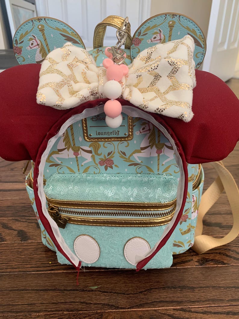 May include: A turquoise blue and gold mini backpack with a white and gold bow, red ears, and a white and pink charm. The backpack has a zippered pocket and a gold chain. The backpack has the text "Disney Loungefly" on it.