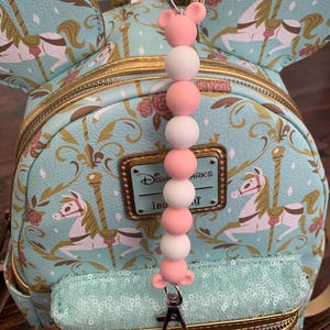 May include: A light blue and gold Disney Parks mini backpack with a carousel print. The backpack has a zippered pocket with sequins and a pink and white beaded keychain with a Mickey Mouse ear charm.