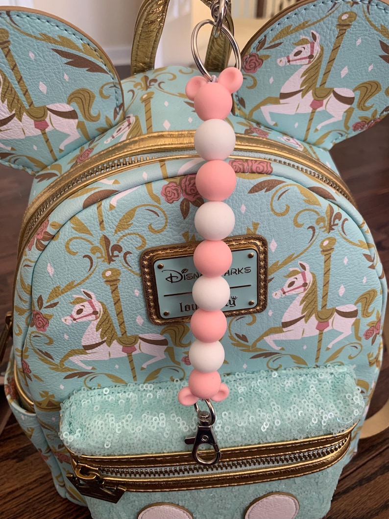 May include: A light blue and gold Disney Parks mini backpack with a carousel print. The backpack has a zippered pocket with sequins and a pink and white beaded keychain with a Mickey Mouse ear charm.