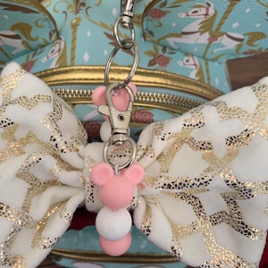 May include: A white and gold bow with a pink and white beaded charm attached to a silver key chain. The charm features a pink Mickey Mouse head.